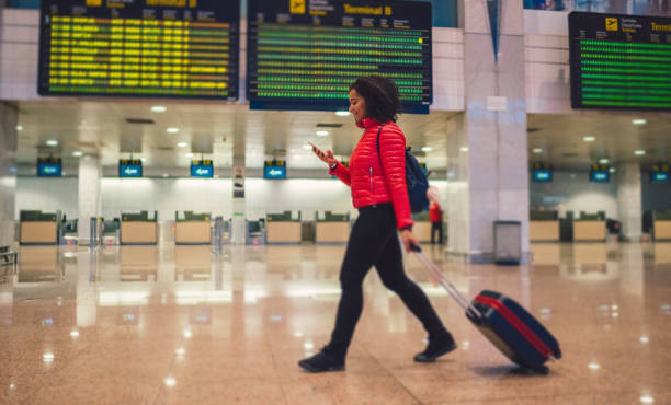 Mixed race woman texting at the airport terminal Young woman with wheeled luggage walking at the airport and texting heathrow airport stock pictures, royalty-free photos & images