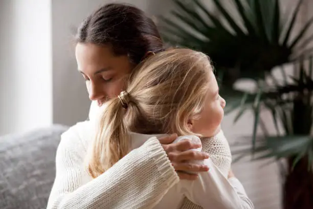 Loving mother hugging cute little girl, young woman embracing adopted child holding tight, sincere warm relationships between mum and daughter cuddling, moms love and care or adoption concept
