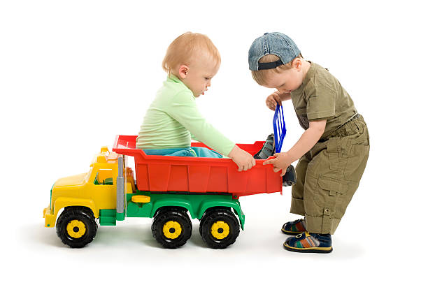 A close-up of a vibrant toy truck sitting in a sandy outdoor play area, partially surrounded by small leaves and grass