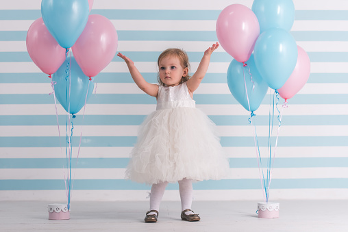 Cute little girl in fluffy white dress is raising hands up while standing near balloons, on light background