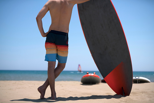 Unrecognizable man surfer standing on the beach and holding his surfboard.