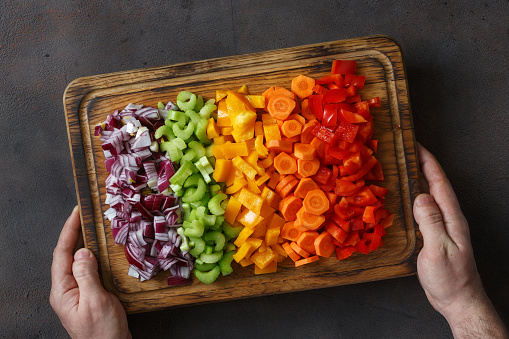 Male hands holding chopped fresh vegetables arranged on cutting board on dark background close up, top view