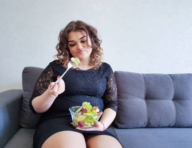 belle fille pleine longueur, assis sur le canapé avec une assiette de salade - overweight women salad frustration photos et images de collection