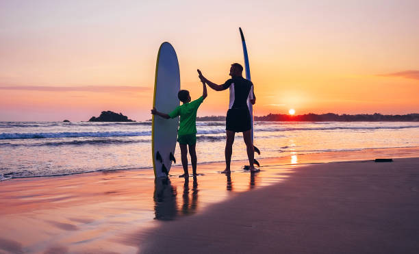 padre e figlio surfisti soggiornano sulla spiaggia al tramonto - surfing beach family father foto e immagini stock