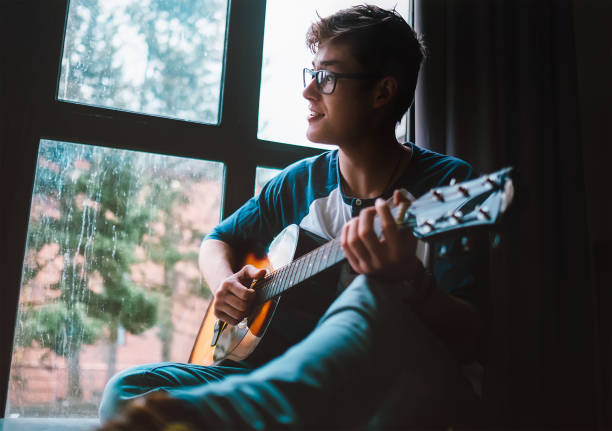 giovane ragazzo che si placca alla chitarra e si siede vicino alla finestra - guitar music guitarist recording studio foto e immagini stock