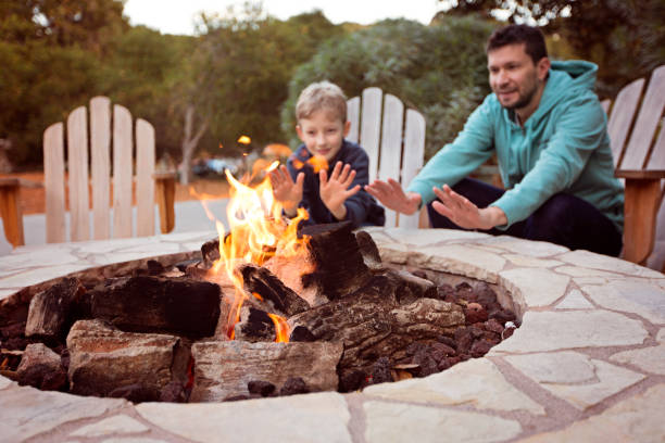 family by firepit view of firepit and happy smiling family of two, father and son, warming their hands by the fire and enjoying time together in the background fire pit photos stock pictures, royalty-free photos & images
