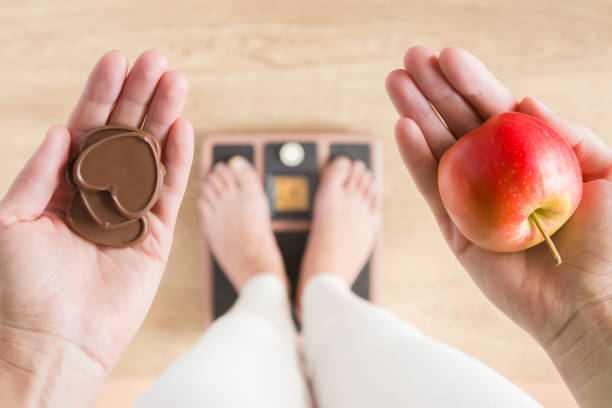 femme debout sur des échelles et la tenue des cœurs de pomme et le chocolat. nouveau départ pour une alimentation saine, minceur du corps, perte de poids. se soucie de corps. dilemme entre fruits ou des bonbons. notion de décision. - fruit variation photos et images de collection