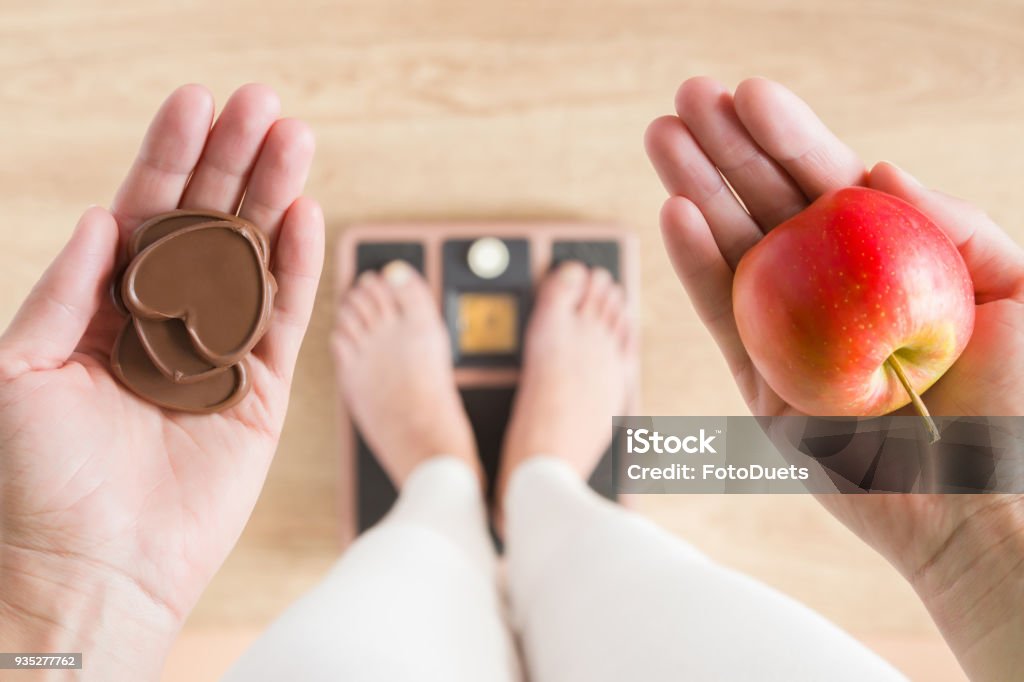 Woman standing on scales and holding apple and chocolate hearts. New start for healthy nutrition, body slimming, weight loss. Cares about body. Dilemma between fruits or sweets. Decision concept. Weight Scale Stock Photo