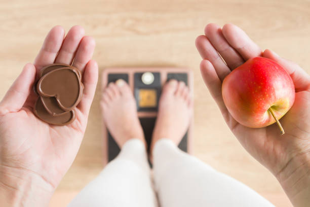 woman standing on scales and holding apple and chocolate hearts. new start for healthy nutrition, body slimming, weight loss. cares about body. dilemma between fruits or sweets. decision concept. - instrument of weight imagens e fotografias de stock