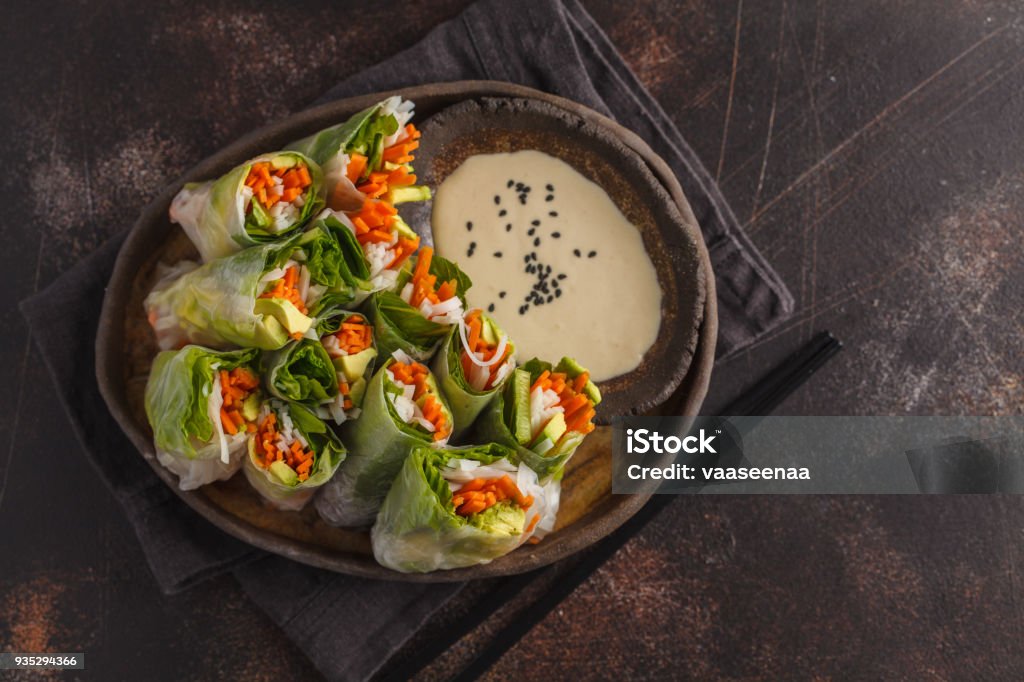 Fresh handmade vegan asian spring rolls with rice noodles, avocado, carrots and tahini dressing on black dish, dark background. Top view, copy space. Fresh handmade vegan asian spring rolls with rice noodles, avocado, carrots and tahini dressing on black dish Lettuce Stock Photo