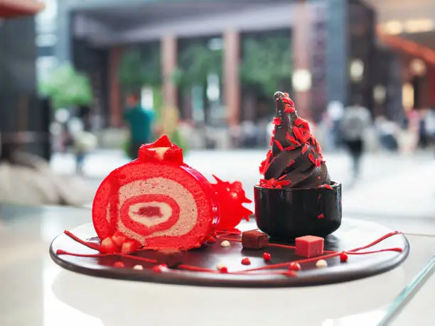 Photo of Black charcoal soft serve ice cream topped with grated strawberry chocolate in black bowl served with strawberry cake roll decorated with chocolate cube and sauce.