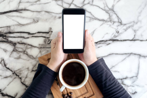 istock Top view mockup image of hands holding black mobile phone with blank white desktop screen and a cup of coffee on table in cafe 935301260