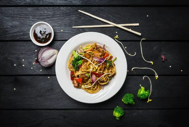 Udon stir fry noodles with meat or chicken and vegetables in a white plate on black wooden background. With chopsticks and sauce. Top view.