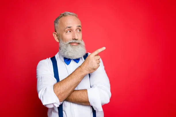 Portrait with copy space of minded, ponder, professional, retro stylist, barber with blue bowtie and suspenders pointing on empty place, product with forefinger, isolated on red background