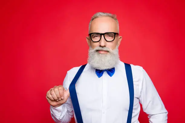 Portrait of elegant, confident macho, old barber, stylist draw off suspender with thumb finger, looking at camera with simper, isolated on red background