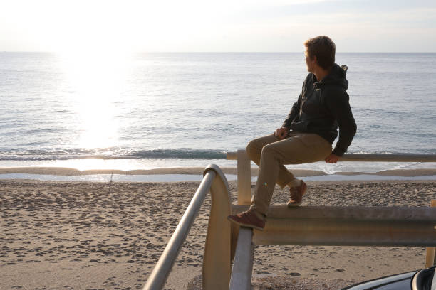 istock Young man sits on railing above car and sea at sunrise 935352354
