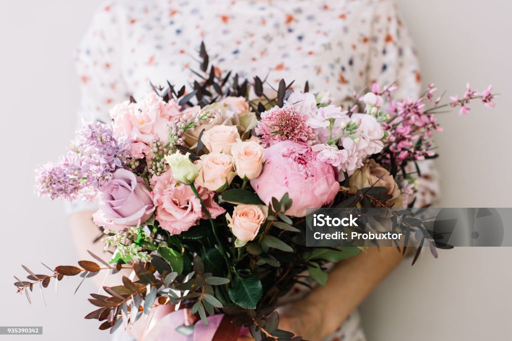 Florist woman holding a beautiful fresh blossoming flower bouquet of peony, roses, lilac, eustoma, mattiola in pink and lavender colors on the grey wall background Women Stock Photo