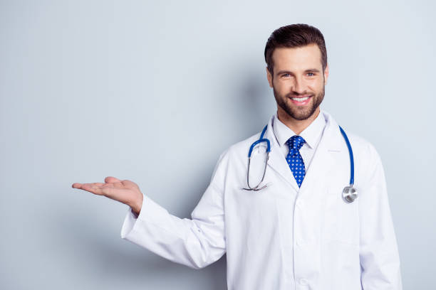 Cheerful handsome bearded smiling guy is holding the copy space. Doc is wearing white uniform and a tie, stands on a light white background