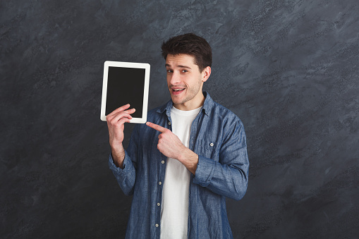 Handsome smiling man pointing at digital tablet with blank screen for commercial advertising at gray studio background, copy space