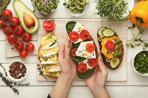 Female hands holding bruschetta with cheese, basil and tomato. Healthy vegetarian sandwiches at kitchen table with various vegetables bowls and greens. Cooking food background, top view