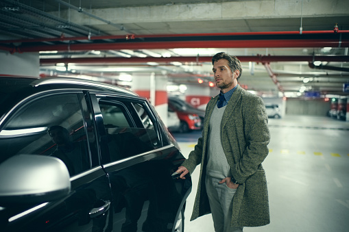 Elegant man standing next to the car parked in the garage and holding hand in pocket.