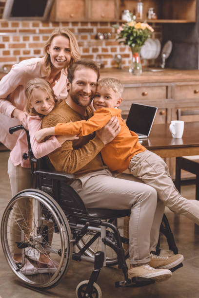 familia feliz con dos niños y el padre en silla de ruedas abrazándose y sonriendo a la cámara en casa - two parent family technology mobility men fotografías e imágenes de stock