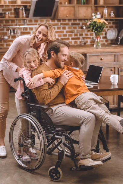 счастливая семья с двумя детьми и отцом в инвалидной коляске обниматься вместе дома - two parent family technology mobility men стоковые фото и изображения