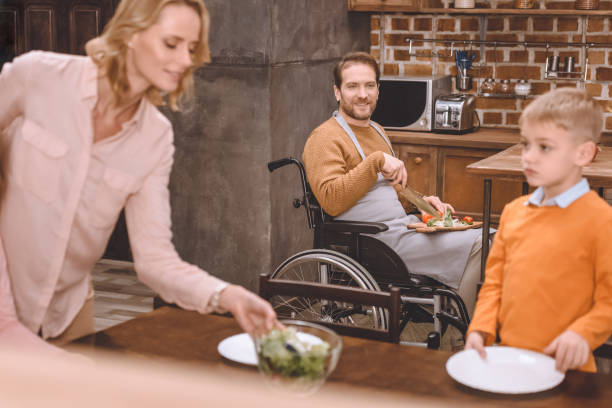 istock mother with son serving table for dinner while father in wheelchair cutting vegetables at home 935480344