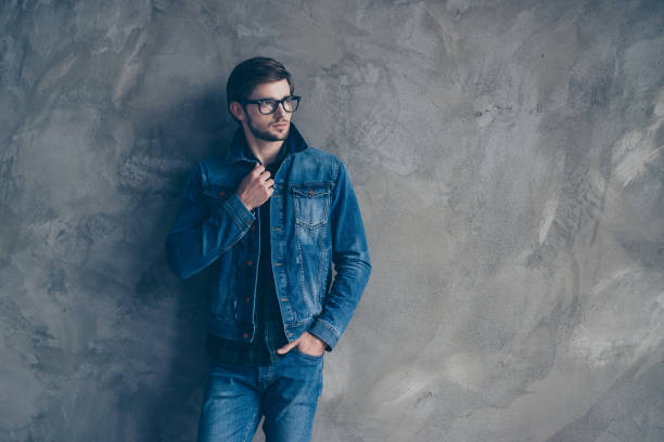 élégant jeune beau mec dans un costume de denim se dresse sur un fond gris et regarde sur le côté - pocket suit glasses jacket photos et images de collection