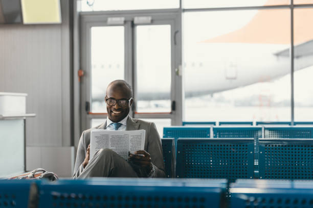 handsome businessman reading newspaper while waiting for flight at airport lobby handsome businessman reading newspaper while waiting for flight at airport lobby newspaper airport reading business person stock pictures, royalty-free photos & images