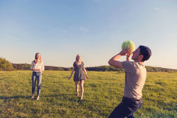 Friends relaxing in park sitting on grass throwing volley ball talking together drinking refreshments
