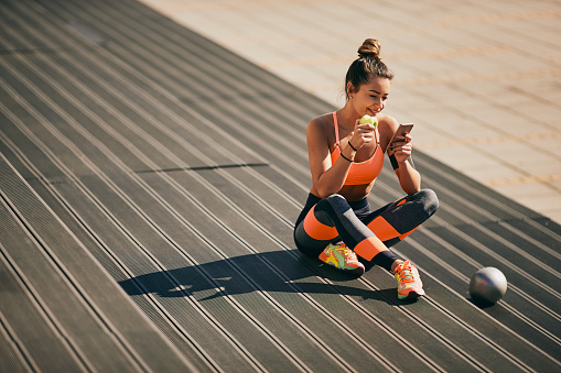 Portrait of girl using smart phone and eating apple while sitting with crossed legs on stairs after exercising.