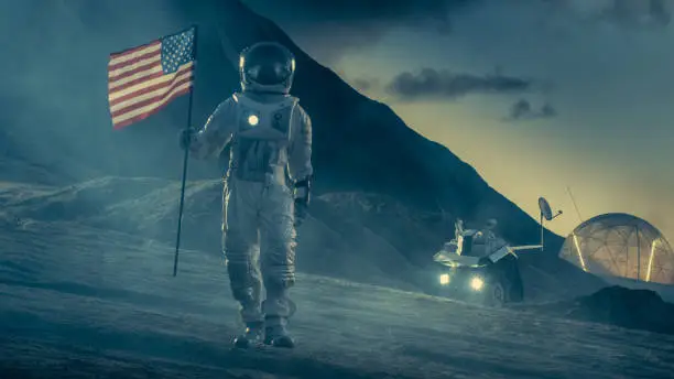 Photo of Strong Astronaut On Alien Planet Walks Through the Storm with a Flag of Unites States of America. In the Background His Research Base/ Station and Rover. Space Travel, Colonisation Theme.