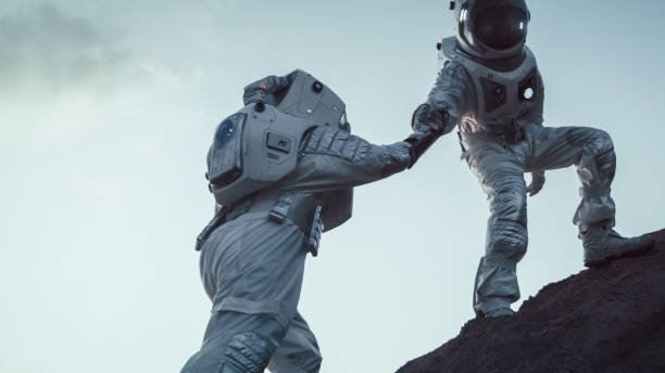 zwei astronauten klettern berghügel helfen einander, oben angekommen. überwindung von schwierigkeiten, wichtiger moment für die menschheit. - super moon stock-fotos und bilder