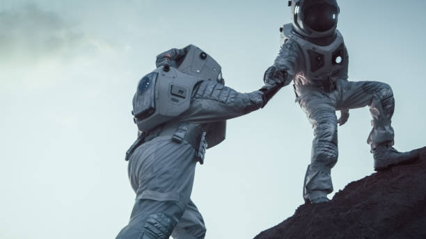 Two Astronauts Climbing Mountain Hill Helping Each Other, Reaching the Top. Overcoming Difficulties, Important Moment for the Human Race. Two Astronauts Climbing Mountain Hill Helping Each Other, Reaching the Top. Overcoming Difficulties, Important Moment for the Human Race. astronaut stock pictures, royalty-free photos & images