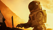 Astronaut in the Space Suit Works on Laptop, Adjusting Rover on a New Alien Red Planet, Presumably Mars. Day Light High-Tech Space Exploration, Mission, Discovering and Colonizing Habitable Planets.