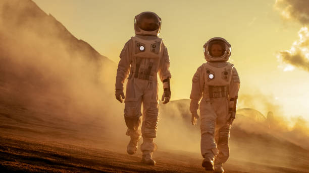 istock Two Astronauts in Space Suits Confidently Walking on Mars, Exploration Expedition on the Planet's Surface. Red Planet Covered in Rocks, Gas and Smoke. Humans Overcoming Difficulties. 935640274