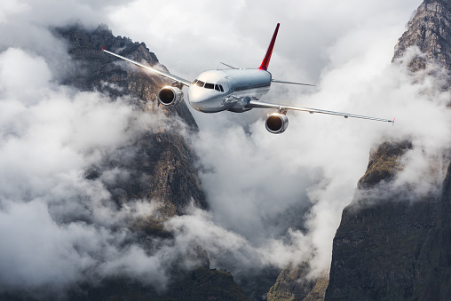 Aircraft, mountains in overcast sky. Airplane is flying in clouds against high mountains. Landscape with airplane, cloudy dramatic sky. Passenger aircraft. Business travel. Commercial plane. Aerial