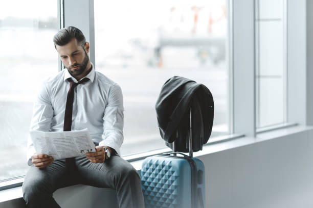 young businessman reading newspaper while waiting for flight at airport lobby young businessman reading newspaper while waiting for flight at airport lobby newspaper airport reading business person stock pictures, royalty-free photos & images