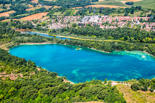 Horizontal vibrant color photography airplane aerial view of vibrant blue lake and wild Ain river, in Auvergne-Rhone-Alpes region in France, Europe. Taken with small town of Priay residential district in background, in summer season with beautiful colors on trees and fields by clear blue sky.