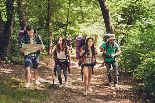 Trekking, camping and wild life concept. Four best friends are hiking in the spring woods, the guy is checking the route on a map, all are excited and joyful
