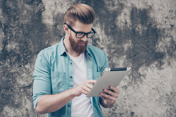 Serious young red bearded man with modern hairstyle in spectacles using digital tablet for reading information near the concrete wall outdoors