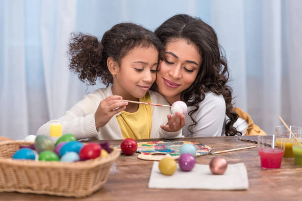 afro americano madre hija abrazando mientras ella pintando huevos de pascua - vacations two generation family holiday easter fotografías e imágenes de stock
