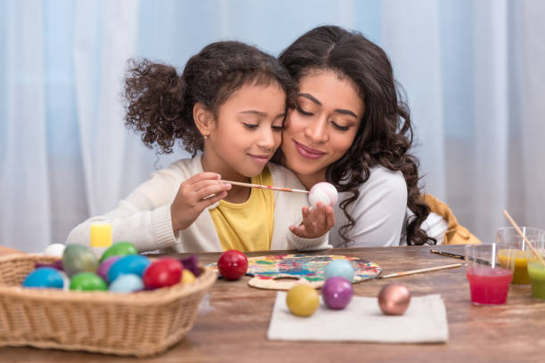 madre afroamericana abbracciando figlia mentre lei dipinge uova di pasqua - easter easter egg paintbrush holding foto e immagini stock