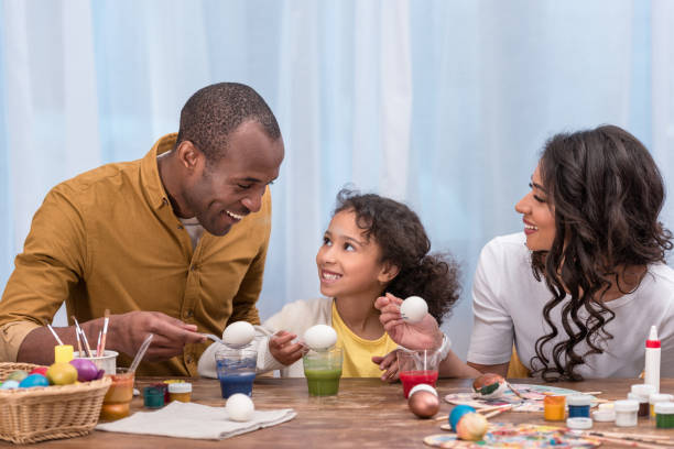 genitori afroamericani e figlia dipingere uova di pasqua e tenendoli sopra bicchieri con vernici - easter easter egg paintbrush holding foto e immagini stock