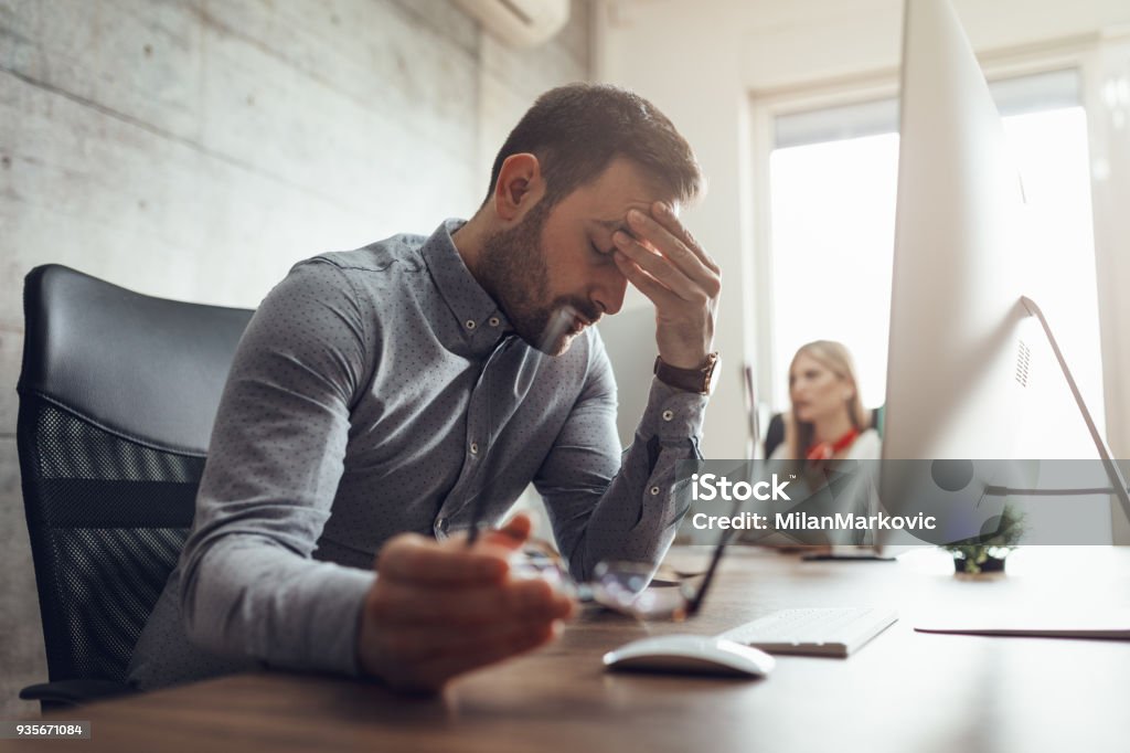 Struggling With Occupational Stress Young handsome frustrated and stressed businessman sitting at the office front a computer and holding head. Emotional Stress Stock Photo