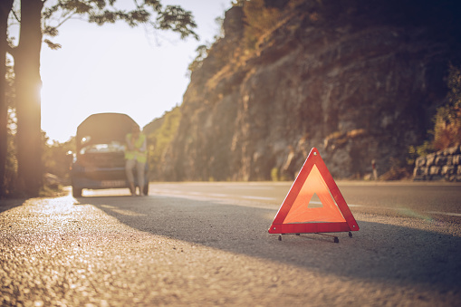 Young woman standing behind warning triangle in front of her broken car on the road
