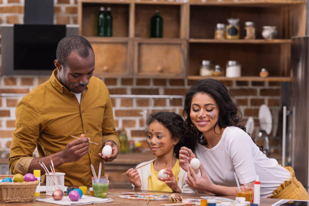 улыбаясь афро-американских родителей и дочь картина пасхальные яйца вместе на кухне - vacations two generation family holiday easter стоковые фото и изображения