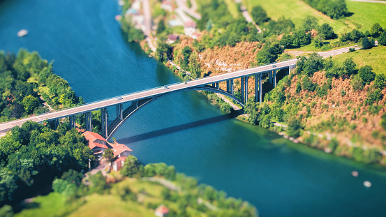 Vibrant color photography, 16x9 composition of aerial view of small cement bridge crossing Ain river, in Auvergne-Rhone-Alpes region in France, Europe. Taken in the end of summer, early in autumn season, with vibrant green trees forest, and wild meadow.