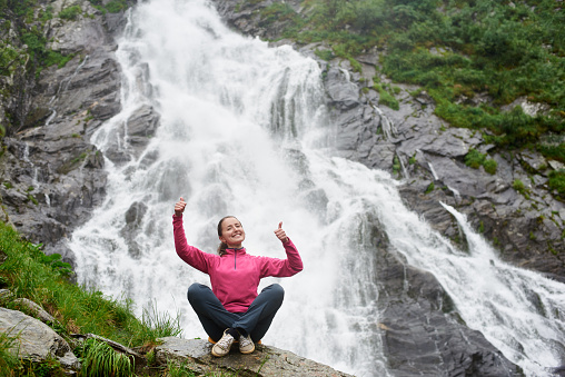Pretty young woman with crossed legs and thumbs up gesture sitting on a large stone at big powerful waterfall. Traveling to the waterfall Balea in Romanian Fagarash mountains.